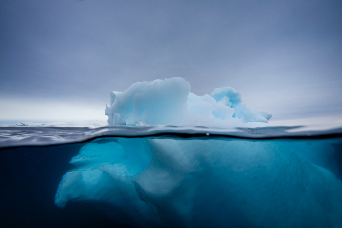 Ein Eisberg vor einem bewölkten Himmel. Auch der Unterwasser liegende Teil ist sichtbar und schimmert türkis.